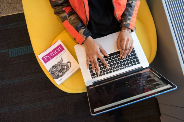 Top view of a skilled developer coding on a laptop, with a Python programming book placed beside them