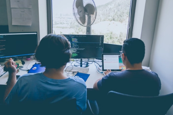 A DevOps and a software engineer collaborating at a workstation with multiple screens displaying code