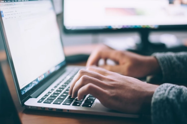 Close-up of a person typing on a laptop keyboard with dual monitors in the background