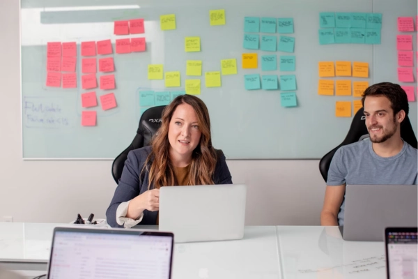 Two people in a meeting room with laptops, discussing ideas in front of a whiteboard covered with sticky notes