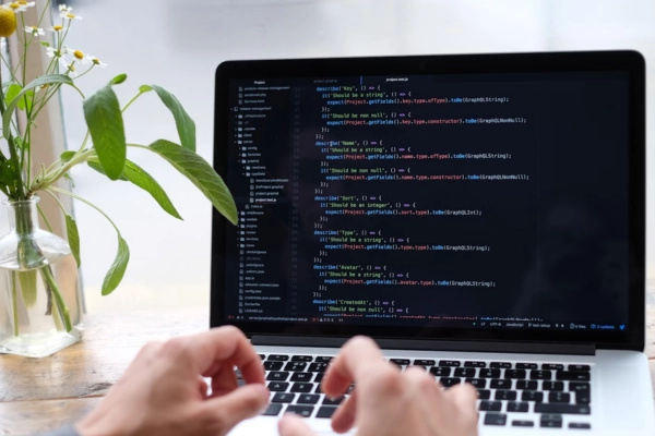 Hands typing on a laptop with lines of code on the screen, placed beside a vase with flowers