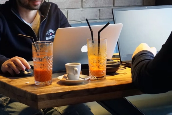 Two people working on laptops at a café table, with orange juice glasses and a coffee cup in the foreground