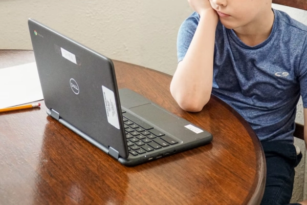 A young boy in a blue shirt using a Dell Chromebook at a wooden table