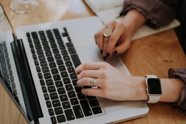 A person wearing a smartwatch and rings typing on a laptop at a wooden table